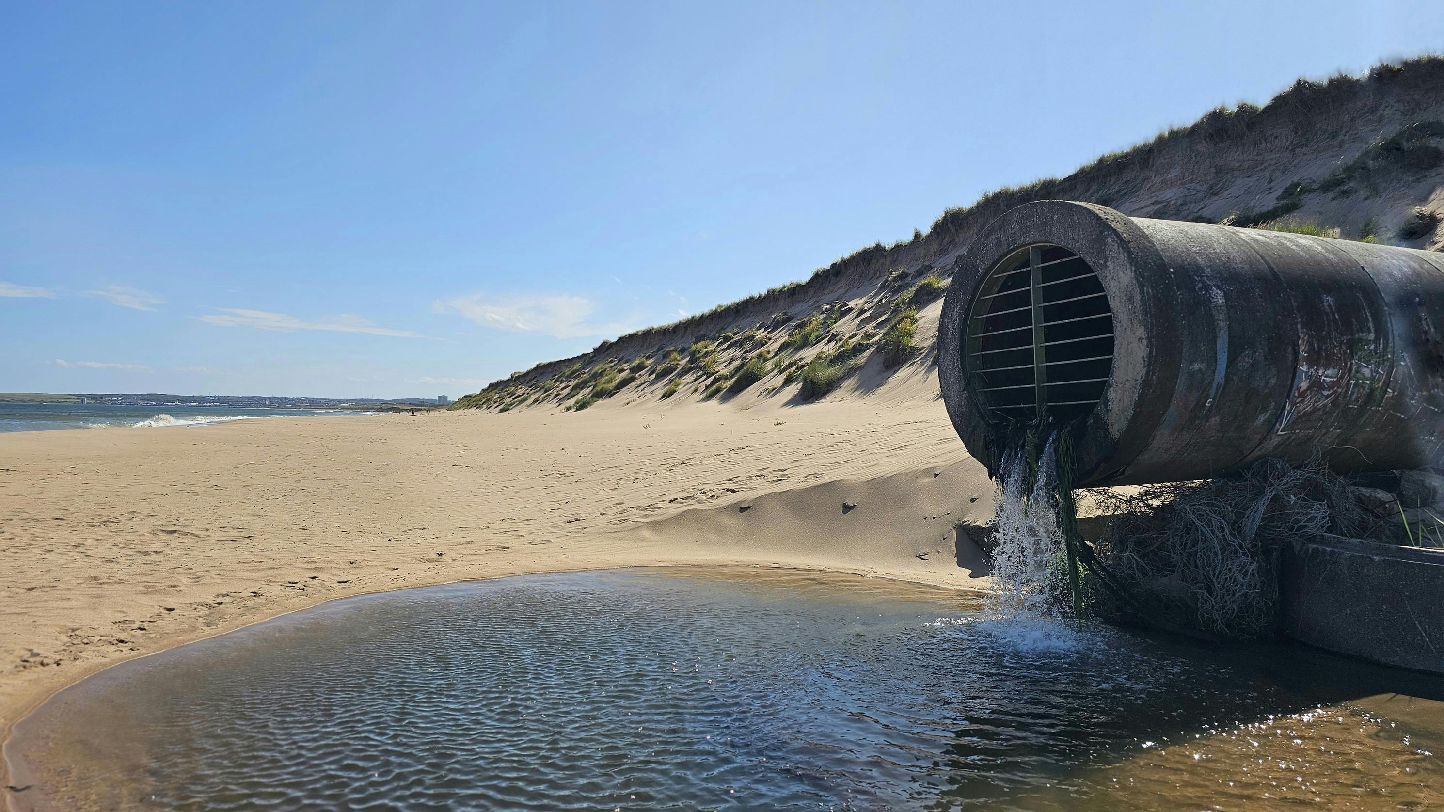 An outflow pipe discharges water onto a sunny beach, punctuating the connection between drainage infrastructure and coastal ecosystems.